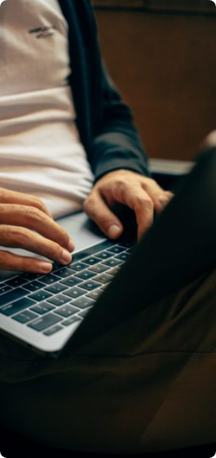 Close-up of a person typing on a laptop, showing their hands on the keyboard. Dressed in a white shirt and brown pants, they work as a partner in de maakindustrie internationaal, with only part of their upper body and face out of frame.